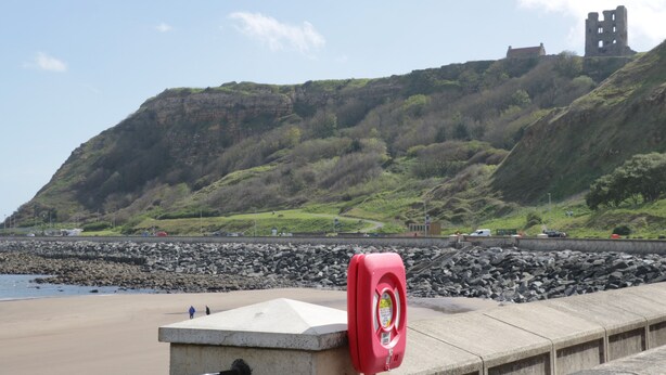 A coastal landscape with a rocky beach, a lifebuoy, and a castle on a hill. - Sanderling (Scarborough)