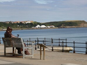 A bench with a bronze statue of a man sitting on it, overlooking a body of water with houses in the background.