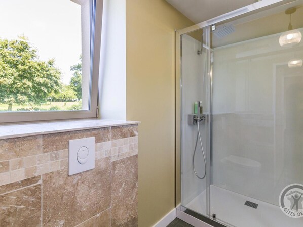 Un baño con cabina de ducha de vidrio, una ventana con vistas a la vegetación y un revestimiento de pared con azulejos.