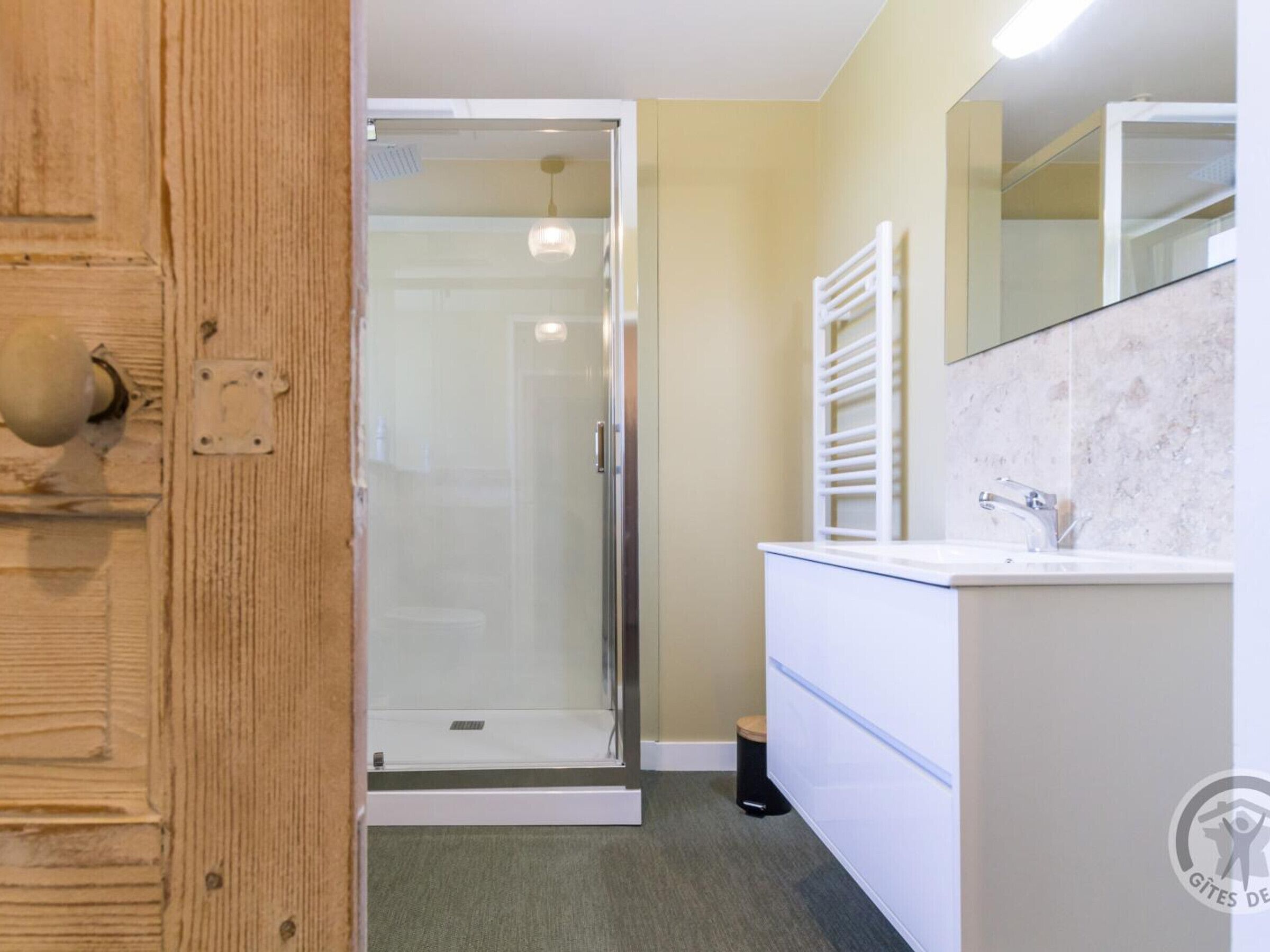 Une salle de bain avec une porte de douche en verre, un lavabo blanc avec un miroir au-dessus, et une porte en bois dotée d’une poignée ronde.