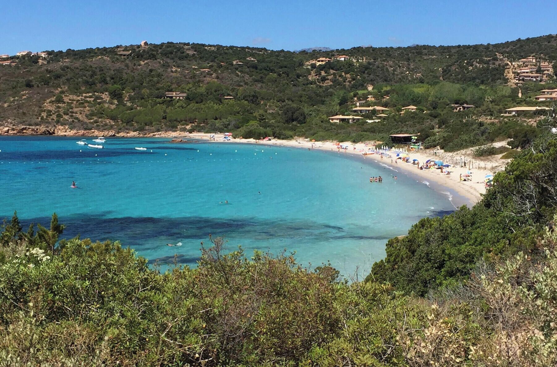 A coastal landscape with a beach, clear blue waters, and buildings on a hillside.
