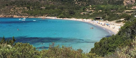 A coastal landscape with a beach, clear blue waters, and buildings on a hillside.