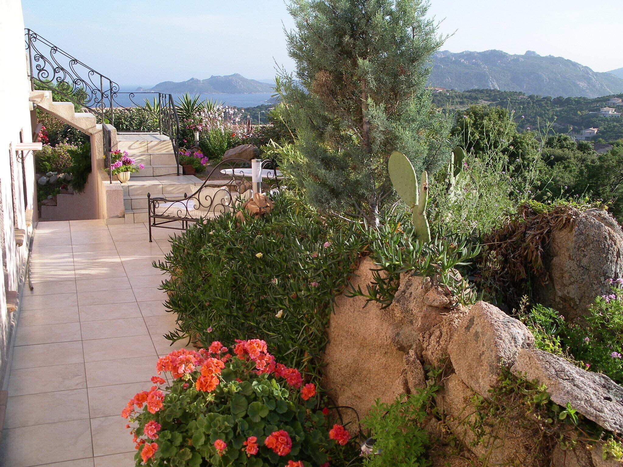 A terrace with a stone staircase, outdoor seating, and a view of the sea and mountains.