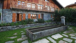 Una casa de madera de dos pisos con base de piedra, contraventanas de madera y un balcón con barandilla de madera.