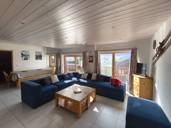 A living room with a sectional sofa, a wooden coffee table, a dining table with chairs, and a view through the glass doors.