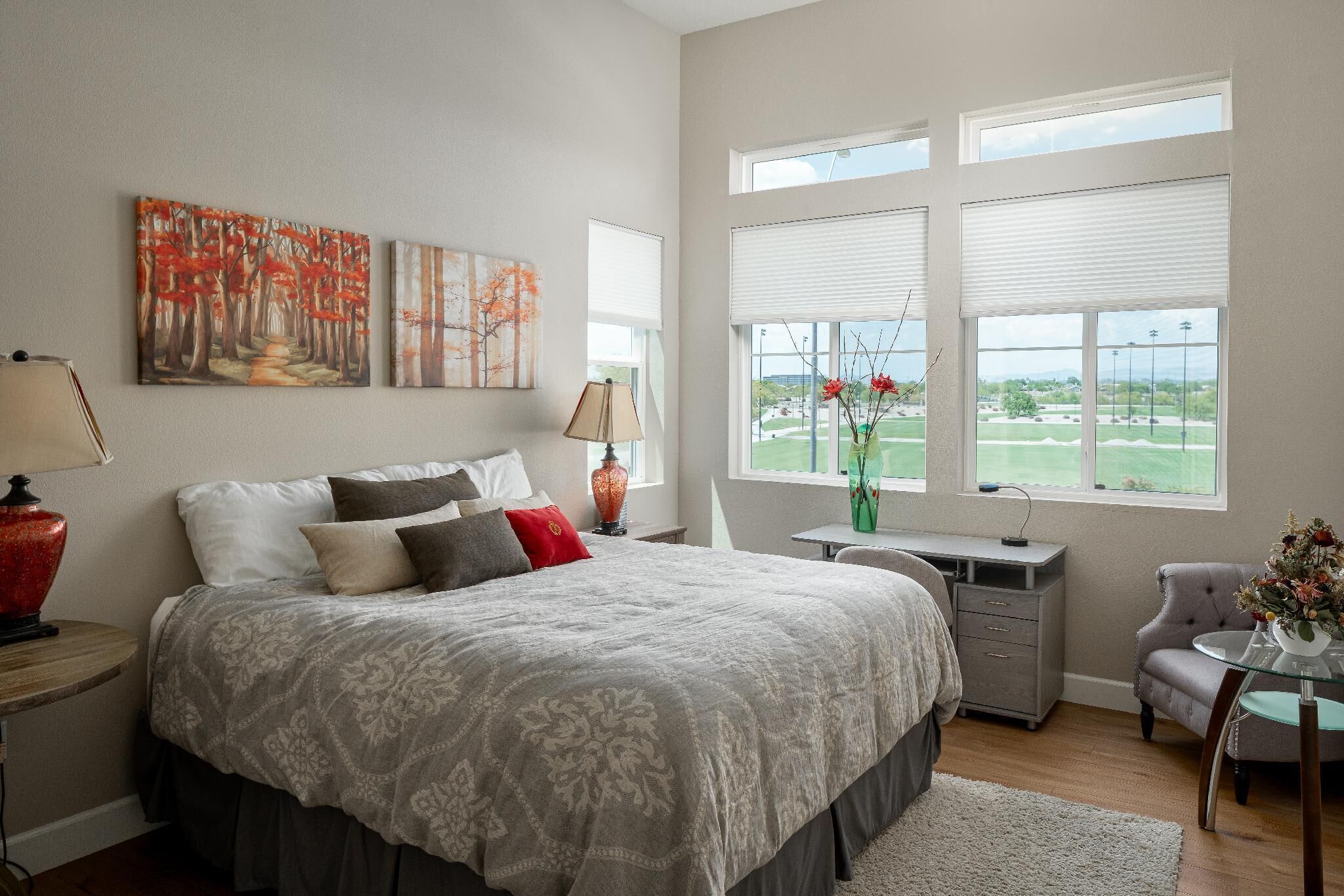 A bedroom with a large bed, a desk, and a chair. There are two windows with blinds, a painting on the wall, and a vase with flowers on the desk.