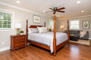 A four-poster bed with a ceiling fan, a wooden nightstand, a gray sofa, and a small table with a plant.