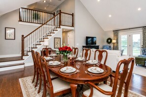 Un coin repas avec une table en bois dressée pour six personnes, un escalier, un salon avec un canapé et un téléviseur.