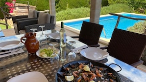 Mesa de jantar com paella de mariscos, copos de vinho e pratos, com vista para uma piscina.