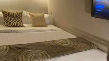 A hotel room with a bed, pillows, a TV mounted on the wall, and a patterned rug.