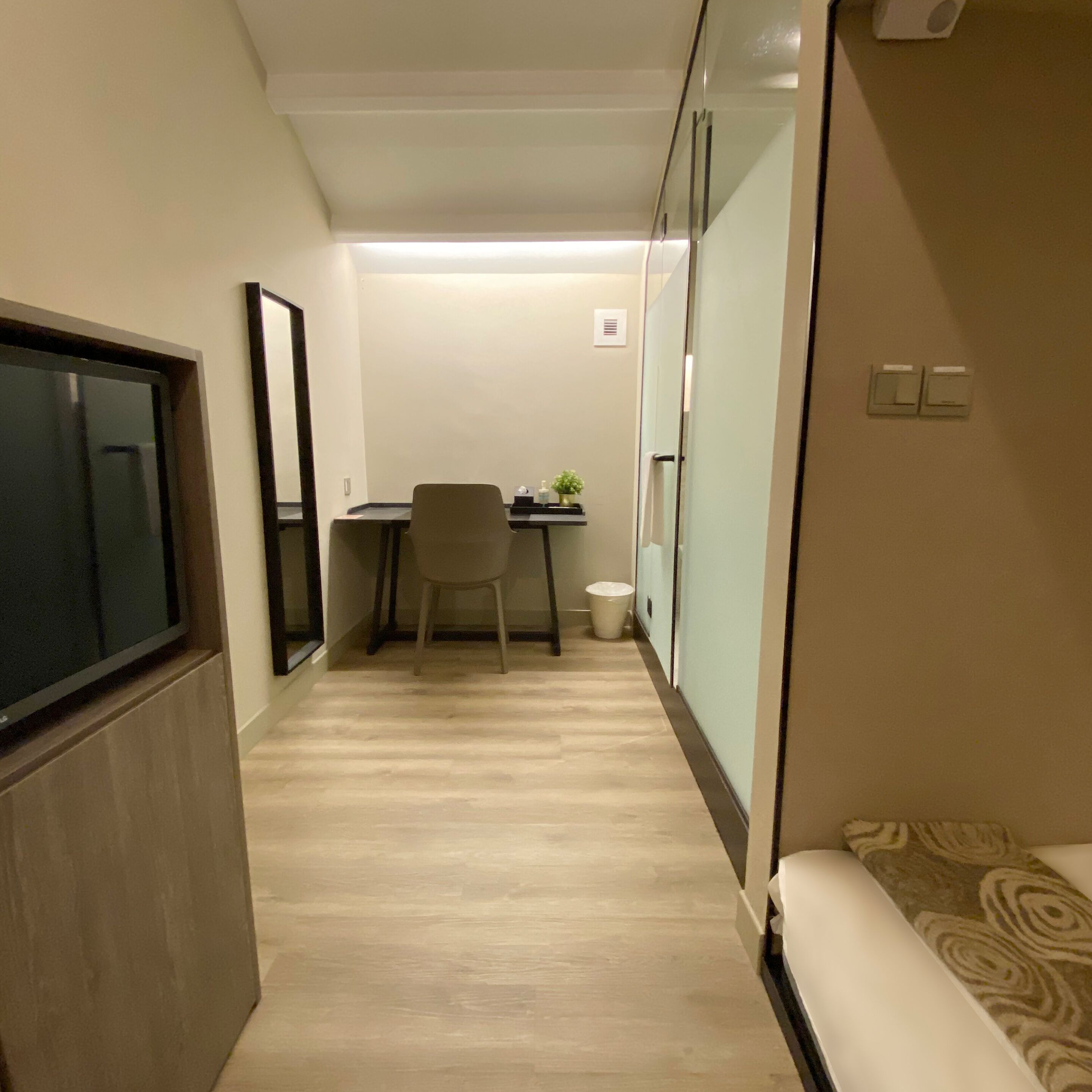 A narrow hallway with a bed on the right, a desk with a chair and a small plant in the center, and a trash can at the end.