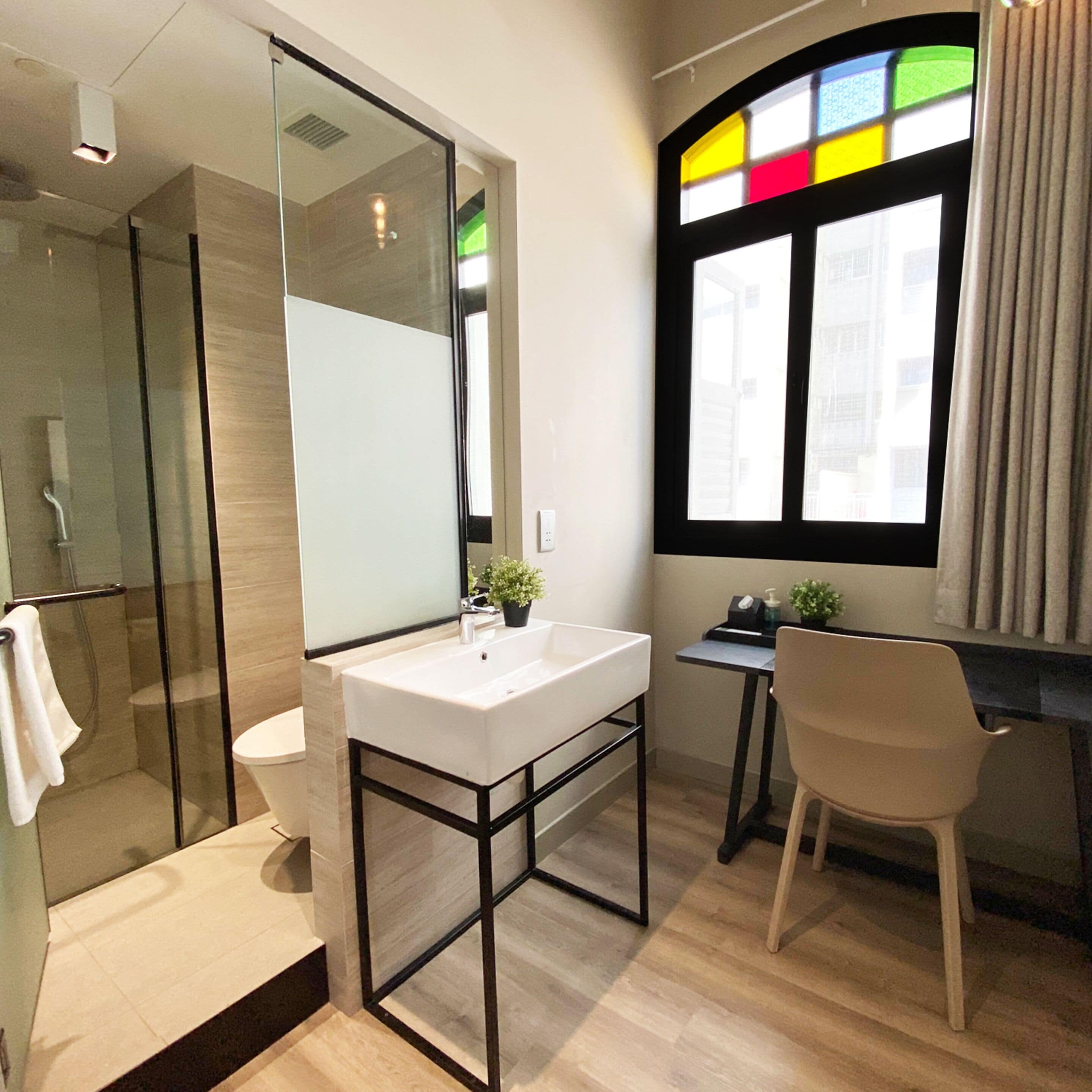A modern bathroom with a glass shower enclosure, a white sink with a black frame, and a window with a stained glass design.