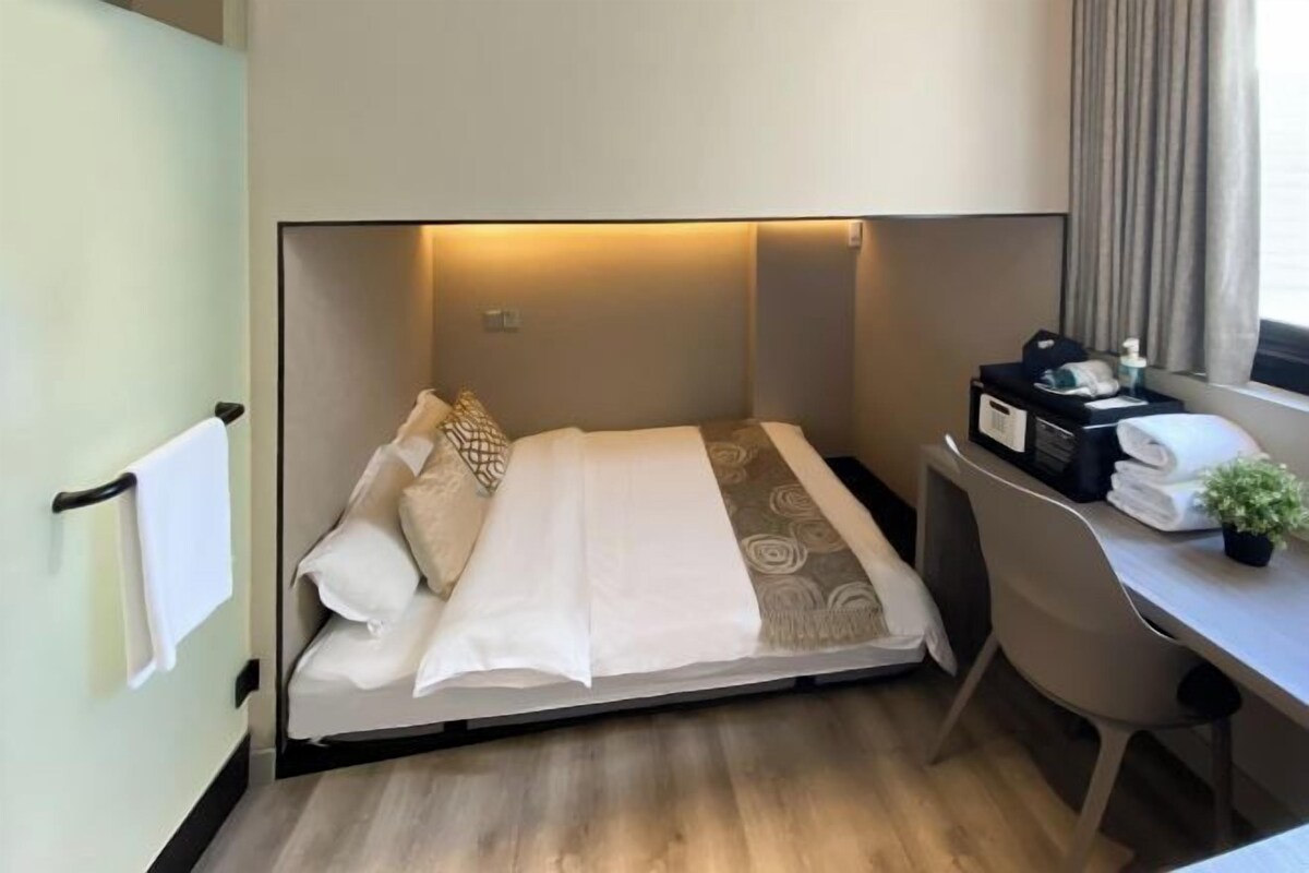 A compact hotel room with a bed, a desk, a chair, a small table with a plant, and a window with curtains.