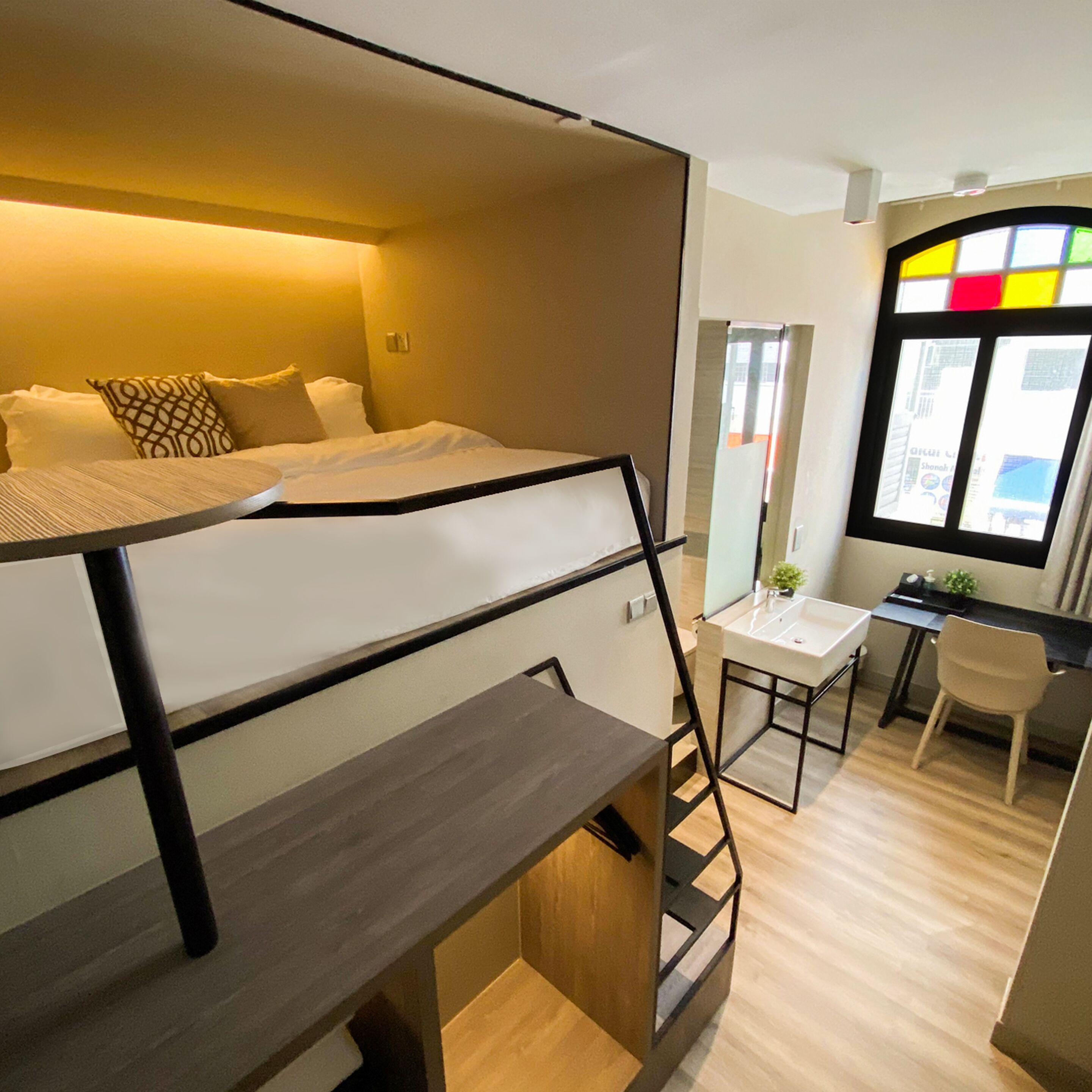 Una habitación de hotel moderna con una cama litera, un escritorio con una silla, una mesita pequeña y una ventana con un diseño de vitral.
