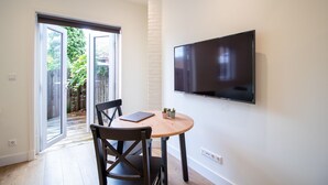 Ein modernes Wohnzimmer mit einem an der Wand montierten Flachbildfernseher, einem Holztisch mit zwei Stühlen und einer Glastür, die in einen Garten führt.