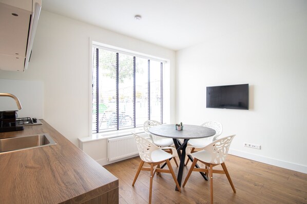 A modern kitchen with a built-in stove, a dining area with a round table and white chairs, and a large window with blinds.