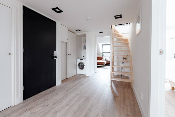 Un couloir avec une porte noire, un lave-linge et un escalier en bois.