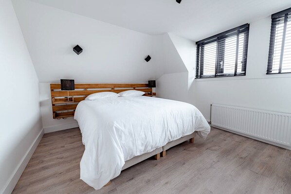 A bedroom with a bed, wooden headboard, white walls, a radiator, and a window with blinds. - Modern and bright apartment (Den Haag)