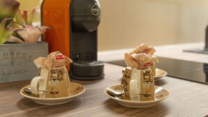 Deux tasses à café ornées d’un motif représentant des grains de café, posées sur une table, une machine à café et un pot contenant des plantes succulentes.