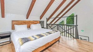A bedroom with a wooden bed, white bedding, and a view of greenery through a balcony railing.