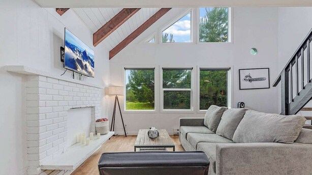 A modern living room with a fireplace, a sofa, a coffee table, and a TV mounted on the wall. There are large windows and a staircase leading to another level.