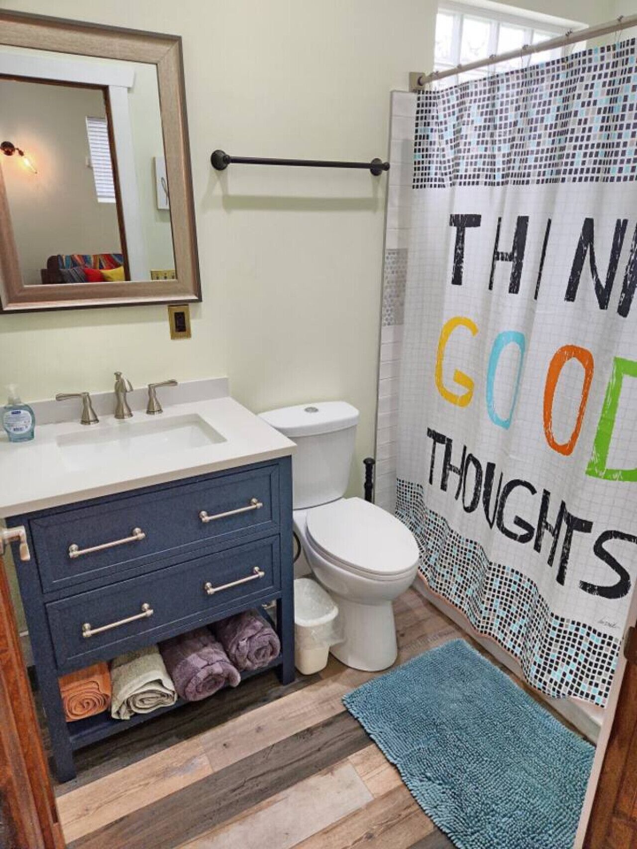 Un baño con un lavamanos blanco, un mueble de color azul oscuro y un inodoro blanco. Se observa una cortina de ducha con la inscripción "THINK GOOD THOUGHTS".
