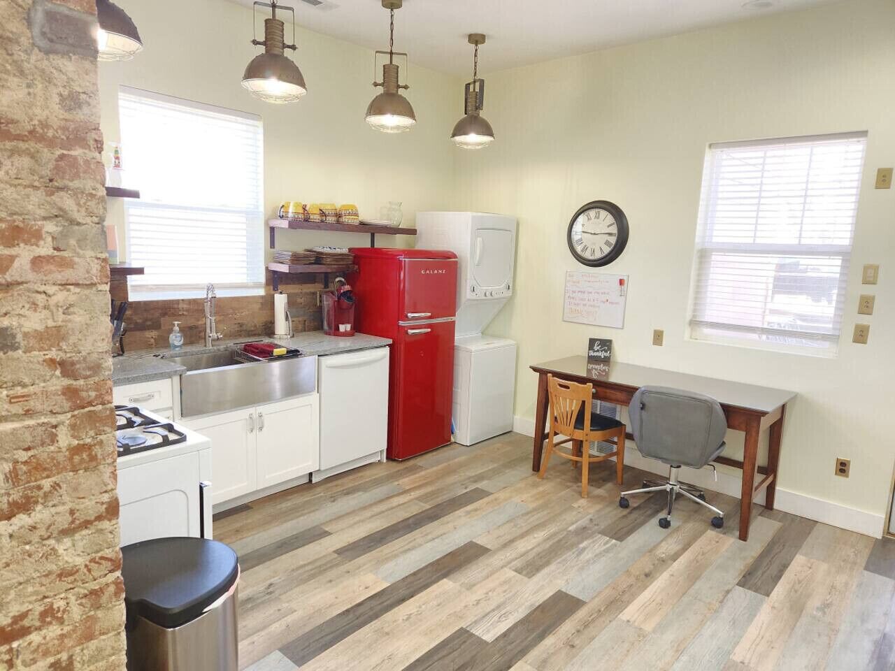 Une cuisine compacte avec des meubles blancs, un réfrigérateur rouge et un coin repas comprenant une table en bois et une chaise de bureau.