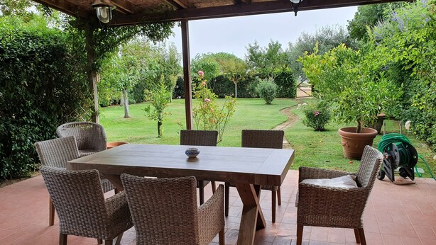 A covered patio with a wooden table and wicker chairs, overlooking a well-maintained garden with various plants and a lawn. - Terraced house a stone's throw from the sea (Capalbio Scalo)
