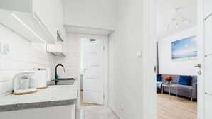 A modern kitchen with white cabinetry, a coffee machine, and a sink. A hallway leads to a living area with a gray sofa and a small table with flowers.