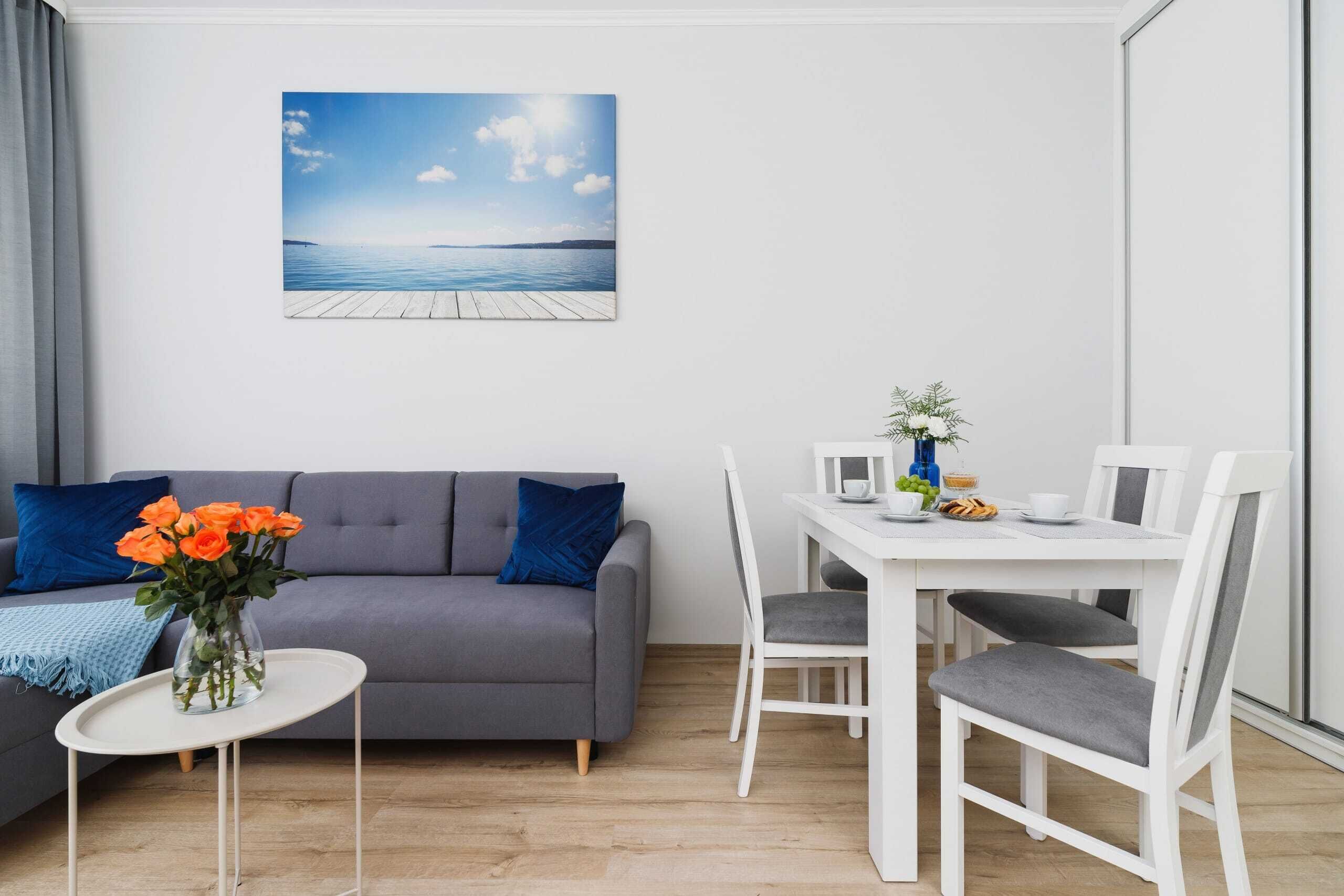 A modern living room with a grey sofa, a small white table, and a dining area with white chairs.