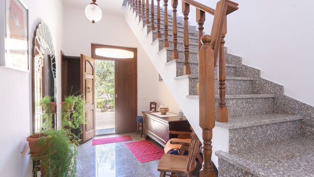 Una escalera con balaustres de madera y peldaños de granito, que conduce a un piso superior. Cerca de la escalera hay una silla de madera y una pequeña mesa.