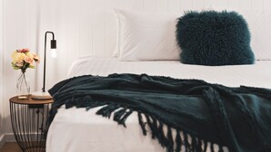 Una cama bien hecha con un edredón verde oscuro, una almohada blanca y una almohada azul mullida sobre una pared blanca. Se ve una pequeña mesa redonda con una lámpara negra y un jarrón con flores.