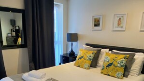 A neatly made bed with white and grey linens, two pillows with floral patterns, and a bedside table with a lamp. - Aspire Apartments (York)