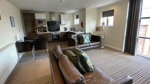 A modern living room with a brown leather sofa, a dining area with a kitchen in the background, and large windows with curtains. - Aspire Apartments (York)