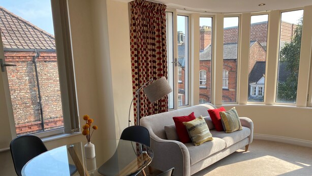 A modern living room with a glass table, black chairs, a beige sofa with red and yellow cushions, and large windows offering a view of brick buildings. - Aspire Apartments (York)