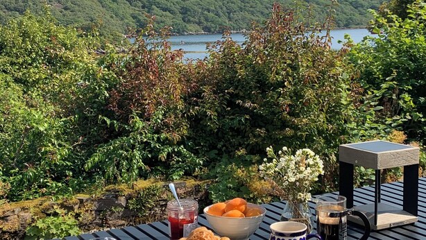 Une table dressée à l’extérieur, avec des croissants, un saladier de fruits, un verre d’eau et une tasse, donnant sur un lac et des collines.