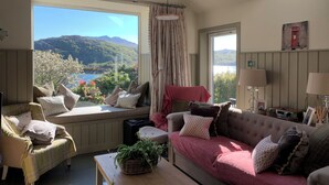 Un salon chaleureux avec vue sur les montagnes et un lac, comprenant un canapé, un fauteuil et une table agrémentée d’une plante.