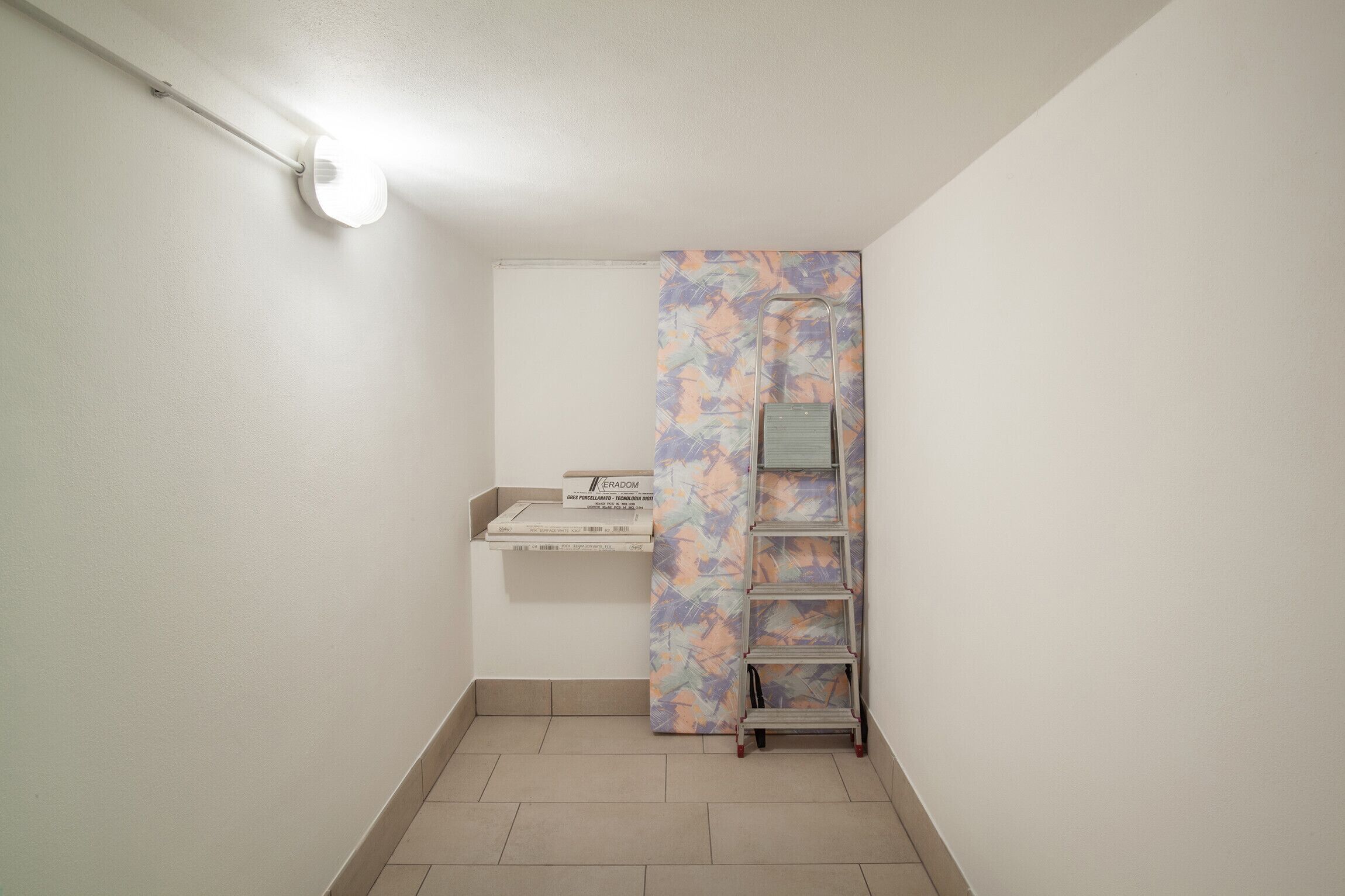 A small, clean room with a white wall, a white ceiling, and a tiled floor. There is a white shelf with a box labeled "ERADOM" and a colorful abstract-patterned curtain. A metal ladder is leaning against the wall.