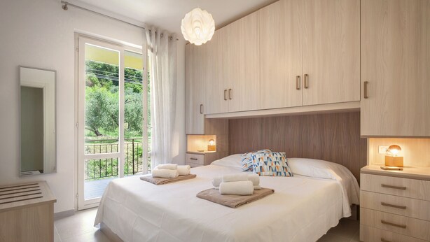 Une chambre moderne avec un grand lit, deux tables de chevet avec des lampes, une armoire en bois et un balcon donnant sur un paysage verdoyant.