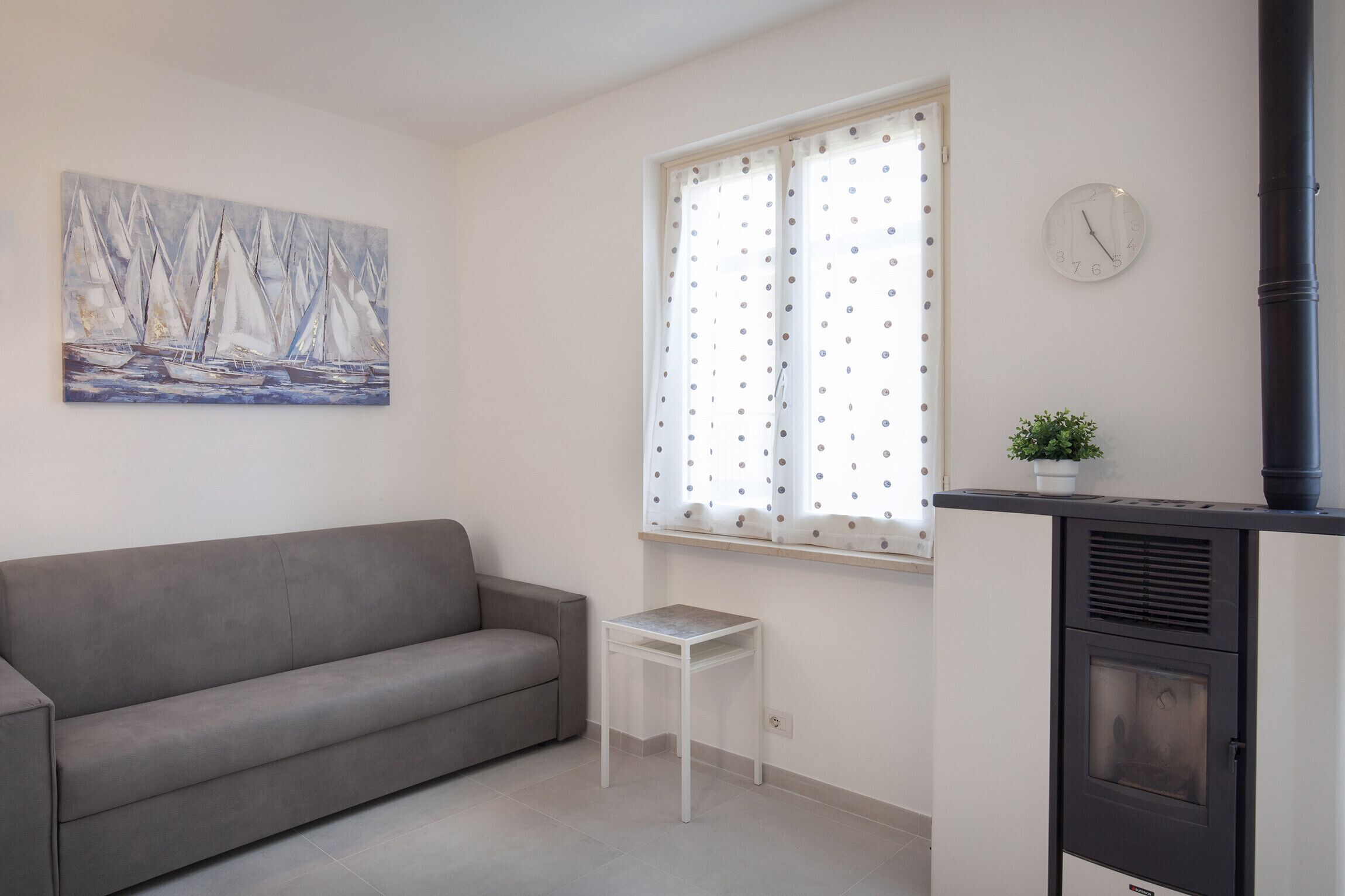 A living room with a grey sofa, a small white table, a window with polka dot curtains, a wall clock, and a black heater.
