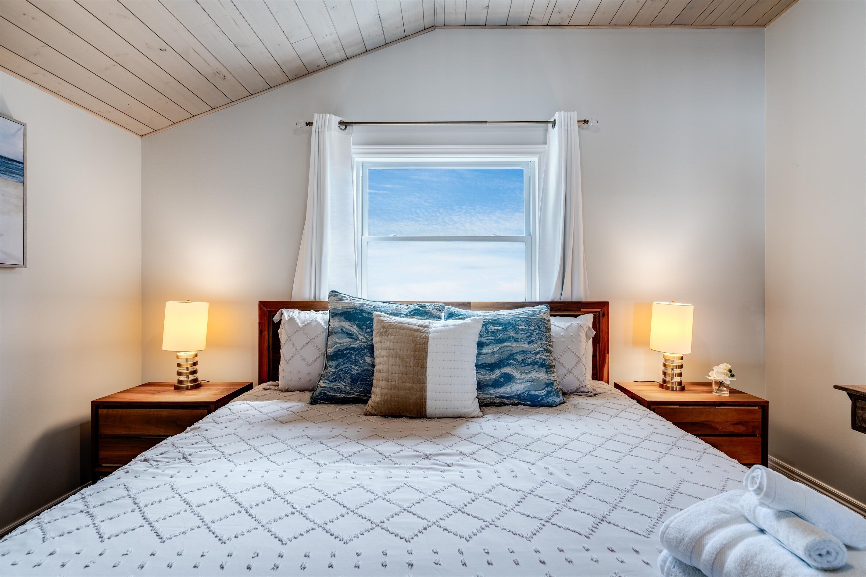 A bedroom with a bed, two bedside tables with lamps, a window with curtains, and a view of the sky.