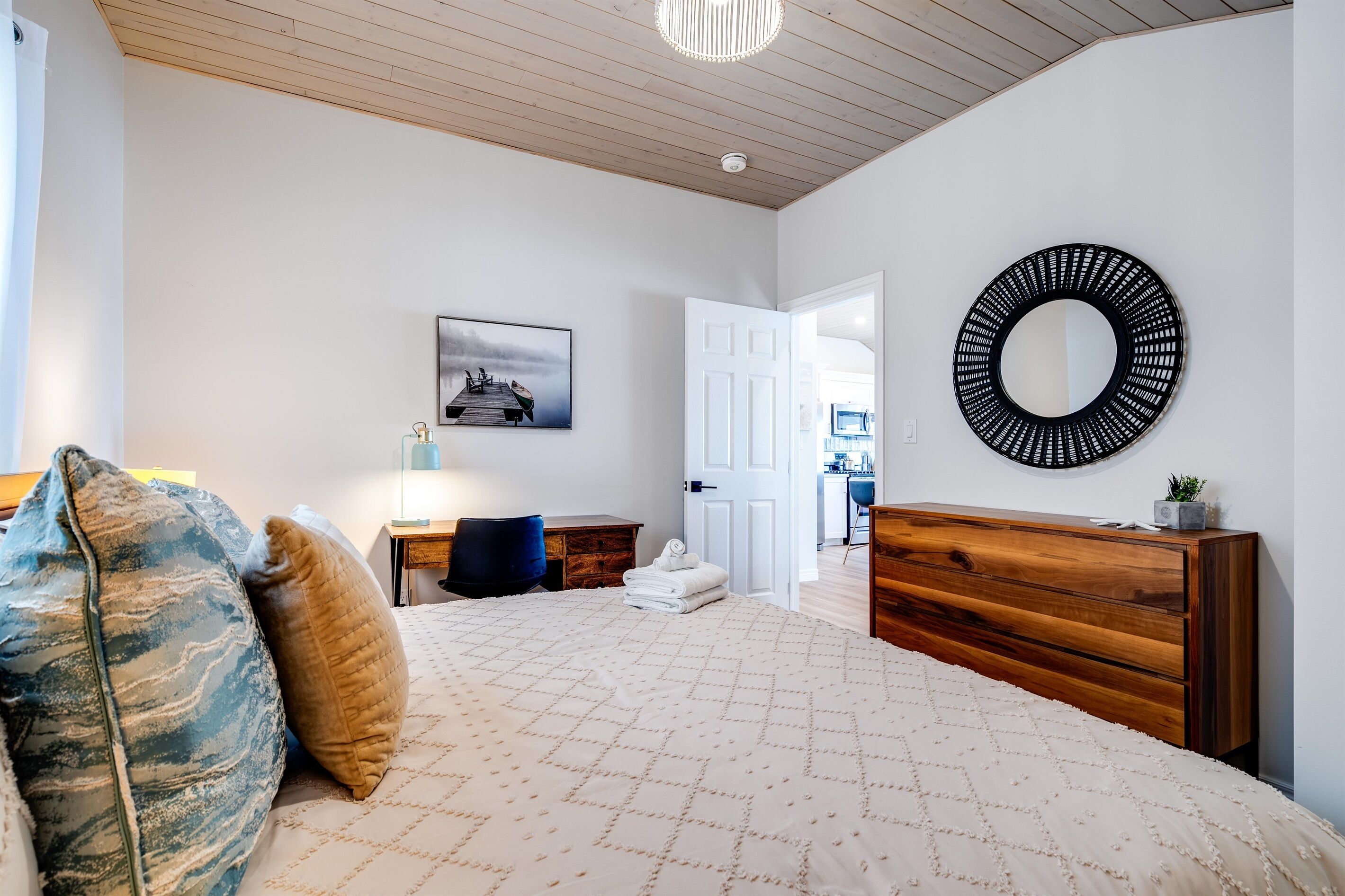 A bedroom with a bed, a wooden dresser, a round mirror, a desk, and a chair.