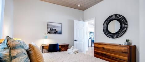 A bedroom with a bed, a wooden dresser, a round mirror, a desk, and a chair.