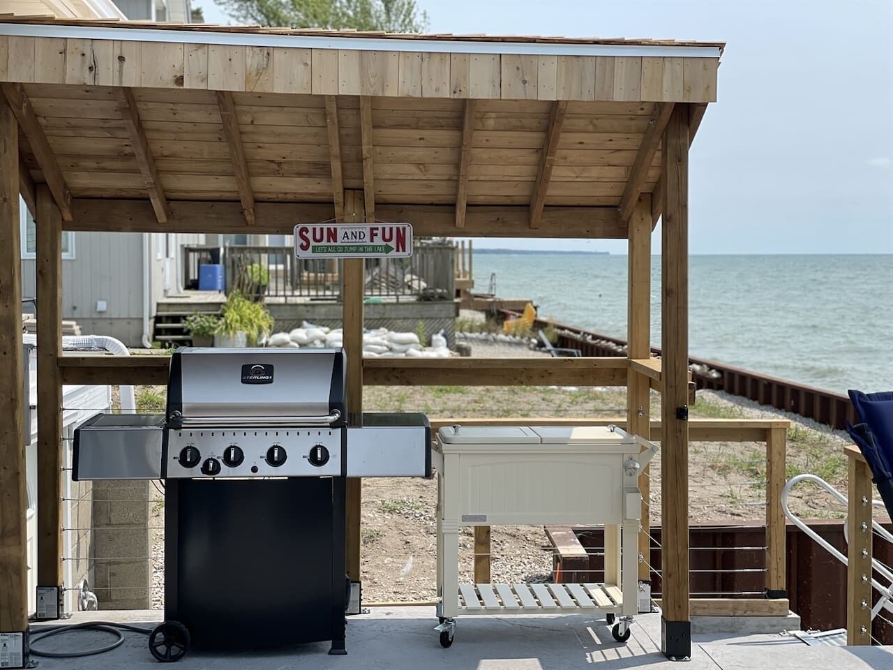 Un espace barbecue extérieur abrité, avec une pancarte indiquant : « SOLEIL ET AMUSEMENT, ALLONS-Y AU LAC », et une vue sur le lac.