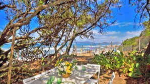 An outdoor dining area with a table set for two, surrounded by greenery and overlooking a beach. - Casa Giannellina (Orbetello)