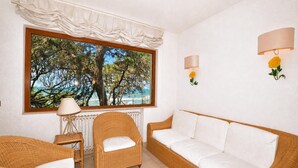 A room with wicker furniture, a large window with a view of trees and the sea, and wall-mounted lamps. - Casa Giannellina (Orbetello)