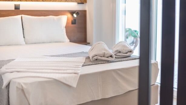 Una cama bien hecha con ropa de cama y toallas blancas, cabecera de madera y vistas al exterior a través de una ventana.