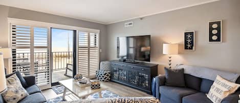 A living room with a sofa, coffee table, and a TV. There is a balcony with sliding doors and a view outside.