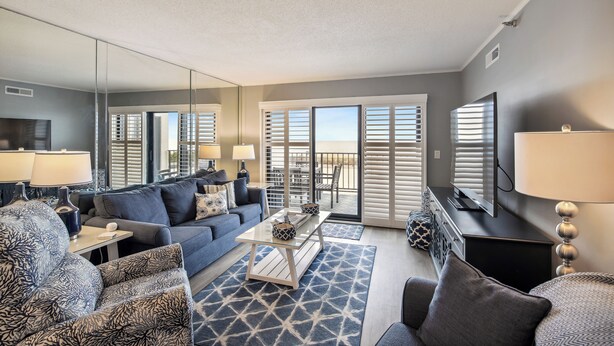 A living room with a sofa, armchairs, a coffee table, and a TV. There is a balcony with sliding doors and a view outside. - XPLORIE ACTIVITIES & LINENS INCLUDED*! PLAZA - 2BR w/Enclosed Den. Amazing ocean views from the balcony! (Ocean City)