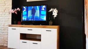 Moderni olohuone, jossa on litteänäyttöinen televisio puisen TV-tason päällä, sen yläpuolella on kello ja molemmin puolin kaksi ruukkuorkideaa.
